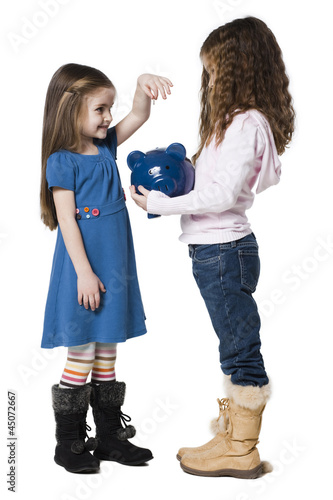 Photo: "Two girls (4-7) putting money in piggy bank, studio shot" "Two girls (4-7) putting money in piggy bank, studio shot"