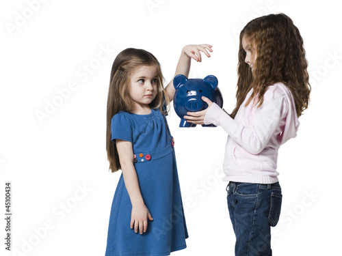 Photo: Studio shot of girl (4-5) inserting money to piggy bank held by sister (6-7) Studio shot of girl (4-5) inserting money to piggy bank held by sister (6-7)