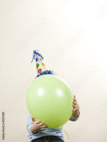Photo: child wearing a party hat holding a balloon in front of his face child wearing a party hat holding a balloon in front of his face