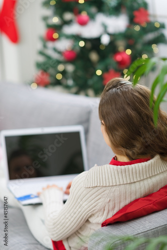 Photo: Woman using laptop near Christmas tree. Rear view Woman using laptop near Christmas tree. Rear view