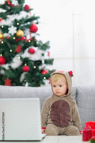 Photo: Baby in Christmas deer costume near Christmas tree looking in la Baby in Christmas deer costume near Christmas tree looking in la