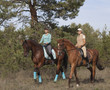 Two smiling girls horseback rides on a trail in the forest.
