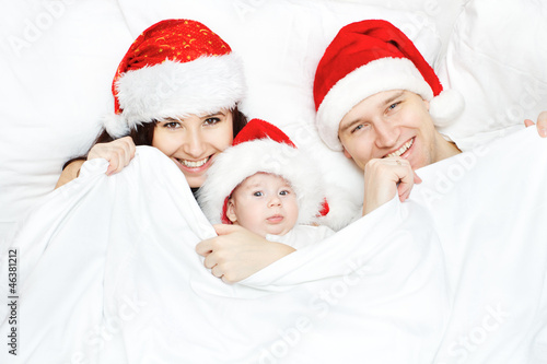 Photo: Christmas family with baby smiling in red hat in white bed Christmas family with baby smiling in red hat in white bed