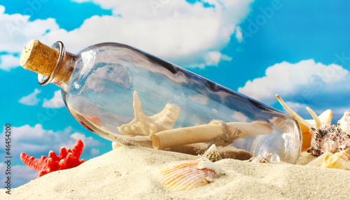 Photo: Glass bottle with note inside on sand, on blue sky background Glass bottle with note inside on sand, on blue sky background