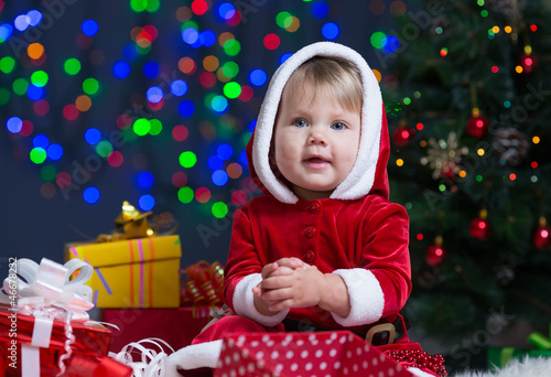 Photo: kid girl dressed as Santa Claus near Christmas tree with gifts kid girl dressed as Santa Claus near Christmas tree with gifts