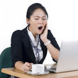Photo: Young business woman yawning at her desk with a cup of coffee Young business woman yawning at her desk with a cup of coffee