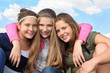 Three happy smiling girls hug at background of sky with clouds.