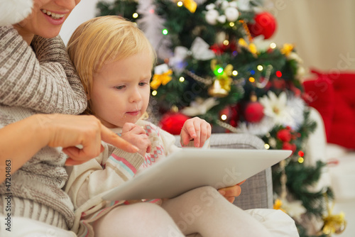Photo: Mother showing baby something in tablet PC near Christmas tree Mother showing baby something in tablet PC near Christmas tree