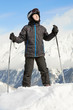 Boy in ski suit stands leaning on ski poles and looking upward,