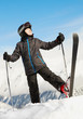 Boy in ski suit stands rising up one leg with ski and looking up