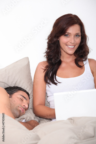Photo: woman having fun on her laptop while her husband is sleeping woman having fun on her laptop while her husband is sleeping
