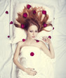Beautiful red-haired girl in bed with rose petal. Studio shot.