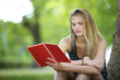 Young woman sitting next to a tree reading from a notebook.