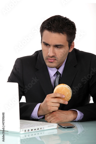 Photo: Man sat at his desk eating hamburger Man sat at his desk eating hamburger