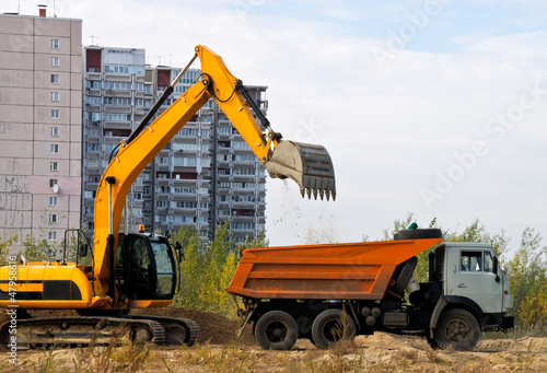 Photo: Excavator loads the ground in a truck on a background of houses Excavator loads the ground in a truck on a background of houses