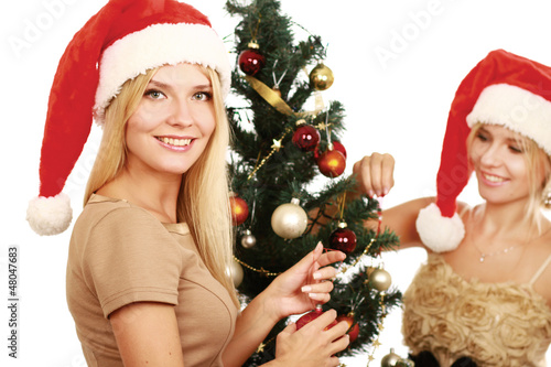Photo: Two girlfriends in santa hat and near Christmas tree Two girlfriends in santa hat and near Christmas tree