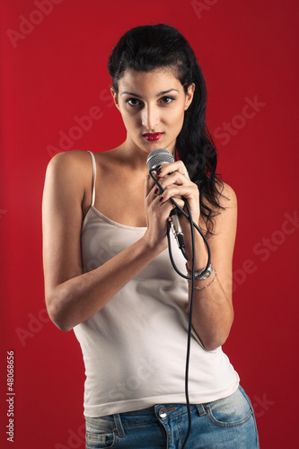 Photo: Beautiful girl singing with microphone against red background. Beautiful girl singing with microphone against red background.