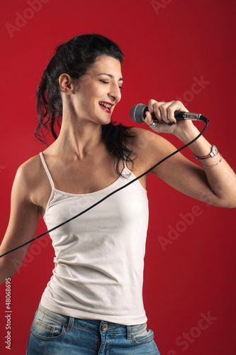 Photo: Beautiful girl singing with microphone against red background. Beautiful girl singing with microphone against red background.