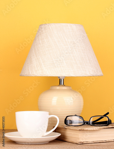 Photo: table lamp with cup, book and glasses on yellow background table lamp with cup, book and glasses on yellow background