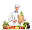 Chef holding a tomato and bag behind a table full of fruits and