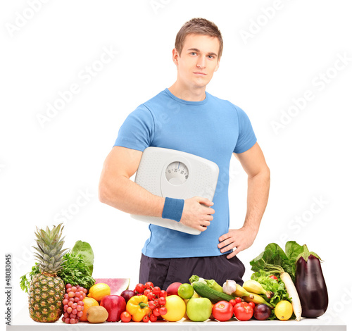 Photo: Male athlete holding a weight scale behind a table full of food Male athlete holding a weight scale behind a table full of food