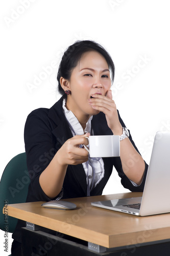 Photo: Young business woman yawning at her desk with a cup of coffee Young business woman yawning at her desk with a cup of coffee