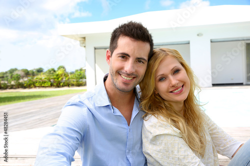 Photo: Cheerful young couple sitting in front of modern house Cheerful young couple sitting in front of modern house
