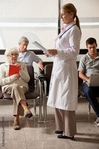 Photo: Doctor Reading File With People In Lobby Doctor Reading File With People In Lobby
