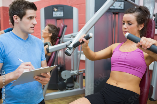 Photo: Trainer noting progress of woman using weights machine Trainer noting progress of woman using weights machine