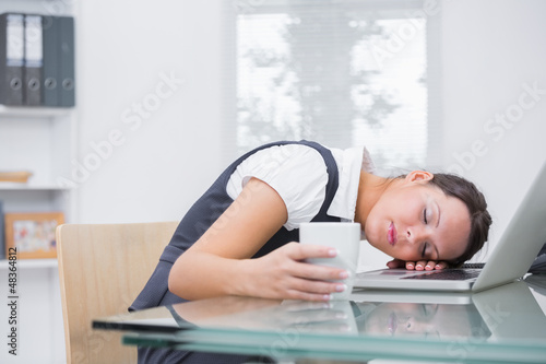 Photo: Business woman with coffee cup resting head on laptop at office Business woman with coffee cup resting head on laptop at office