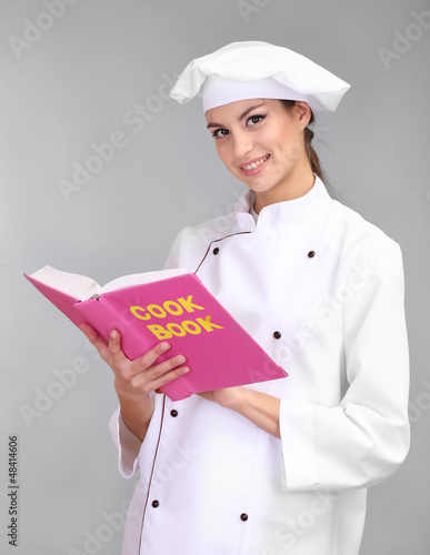 Photo: Portrait of young woman chef with cookbook on grey background Portrait of young woman chef with cookbook on grey background