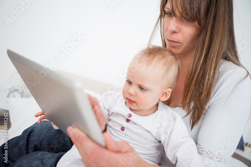 Photo: Serious woman holding her baby while using a tablet pc Serious woman holding her baby while using a tablet pc