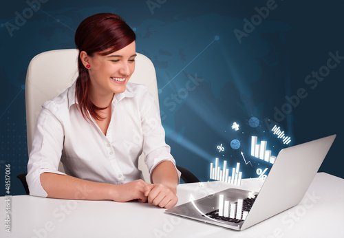 Photo: Pretty woman sitting at desk and typing on laptop with diagrams Pretty woman sitting at desk and typing on laptop with diagrams