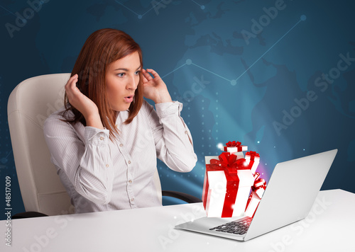 Photo: Pretty lady sitting at desk and typing on laptop with present bo Pretty lady sitting at desk and typing on laptop with present bo