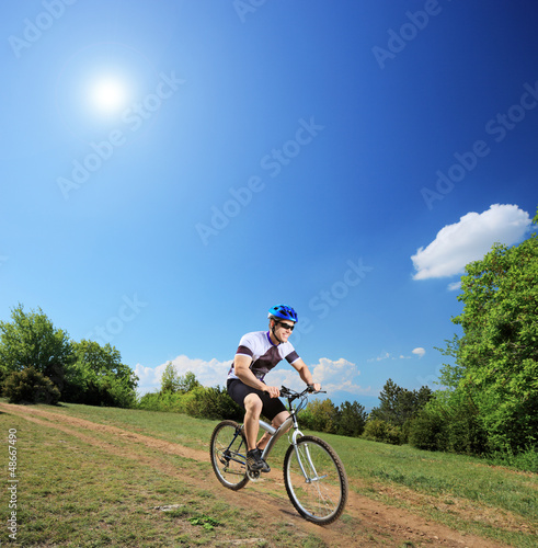 Photo: Male bicyclist riding a mountain bike down hill on a sunny day Male bicyclist riding a mountain bike down hill on a sunny day