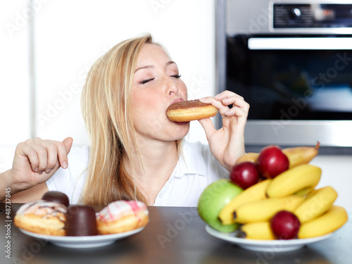 Photo: Pretty woman choosing between fruits and sweets, enjoys a donut Pretty woman choosing between fruits and sweets, enjoys a donut