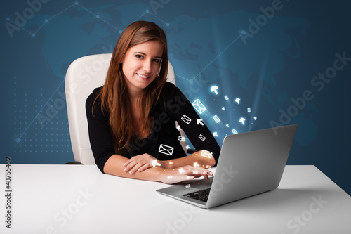 Photo: Young lady sitting at dest and typing on laptop with message ico Young lady sitting at dest and typing on laptop with message ico