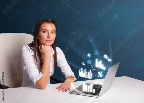 Photo: Pretty woman sitting at desk and typing on laptop with diagrams Pretty woman sitting at desk and typing on laptop with diagrams