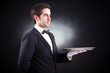 Portrait of a young waiter holding an empty dish on black backgr
