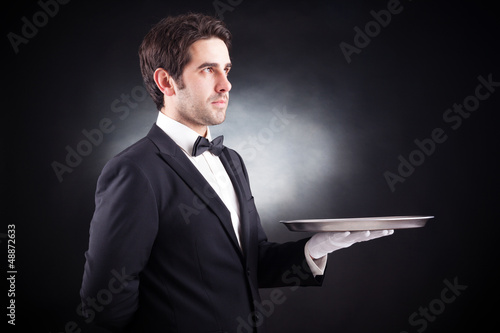 Photo: Portrait of a young waiter holding an empty dish on black backgr Portrait of a young waiter holding an empty dish on black backgr