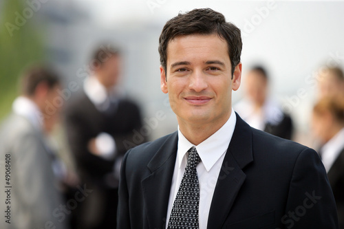 Photo: Businessman stood with colleagues in background Businessman stood with colleagues in background