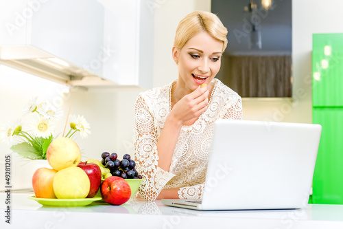 Photo: Beautiful young woman using her laptop computer in the kitchen Beautiful young woman using her laptop computer in the kitchen