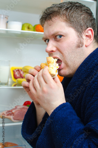 Photo: Handsome man eating piece of cake near open fridge Handsome man eating piece of cake near open fridge