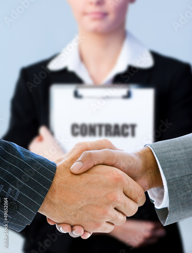 Photo: Businessmen shaking hands with contract behind them Businessmen shaking hands with contract behind them