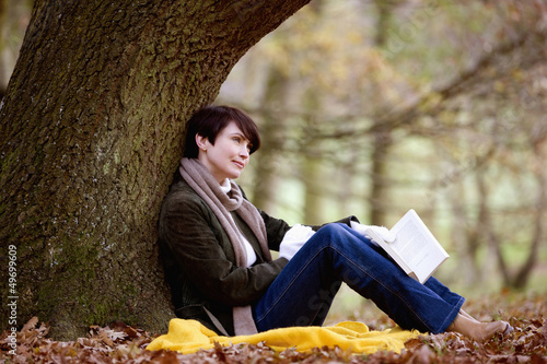 Photo: A woman sitting under a tree in autumn time, holding a book A woman sitting under a tree in autumn time, holding a book