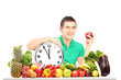 Young man holding a wall clock and apple, sitting on a table ful