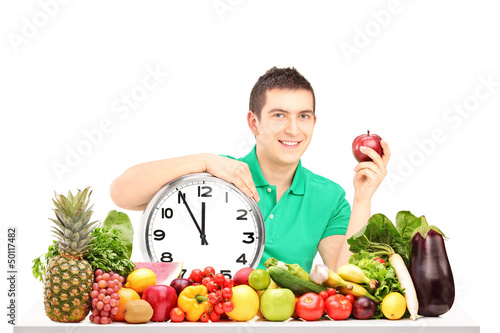 Photo: Young man holding a wall clock and apple, sitting on a table ful Young man holding a wall clock and apple, sitting on a table ful