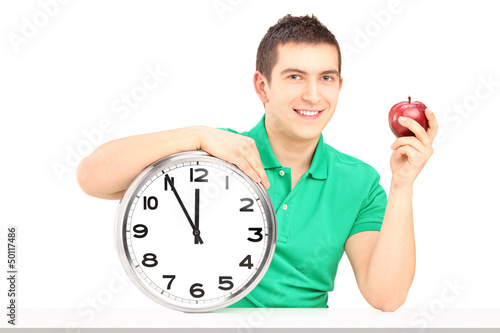 Photo: Young man holding wall clock and red apple on a table Young man holding wall clock and red apple on a table