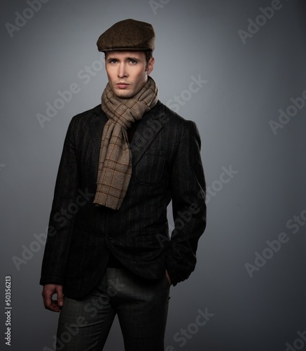 Photo: Young man in brown jacket wearing cap and scarf isolated on grey Young man in brown jacket wearing cap and scarf isolated on grey