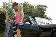 A young couple standing in front of a black sports car kissing